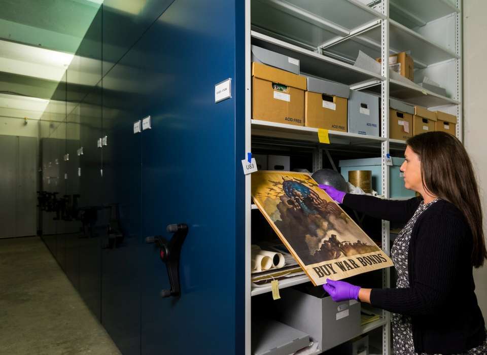 This photograph from the National WWII Museum shows collections boxes stored on Space Saver shelving, in archival boxes, and properly tagged with archival tags. Notice the staff member is also handling the poster with gloves while is fully supported so that it doesn't bend and tear. Image source: https://www.nationalww2museum.org/war/articles/20-years-collecting-wwii-artifacts