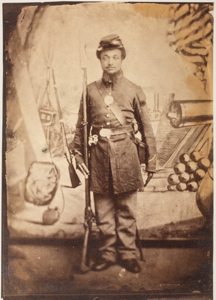 Unidentified African American soldier in uniform posed before a stylized backdrop featuring a canon, other weaponry, a tent, an ironclad gunboat, and an American flag at Benton Barracks in St. Louis, Missouri. He appears to hold a conversion musket with a fixed bayonet, possibly a prop.